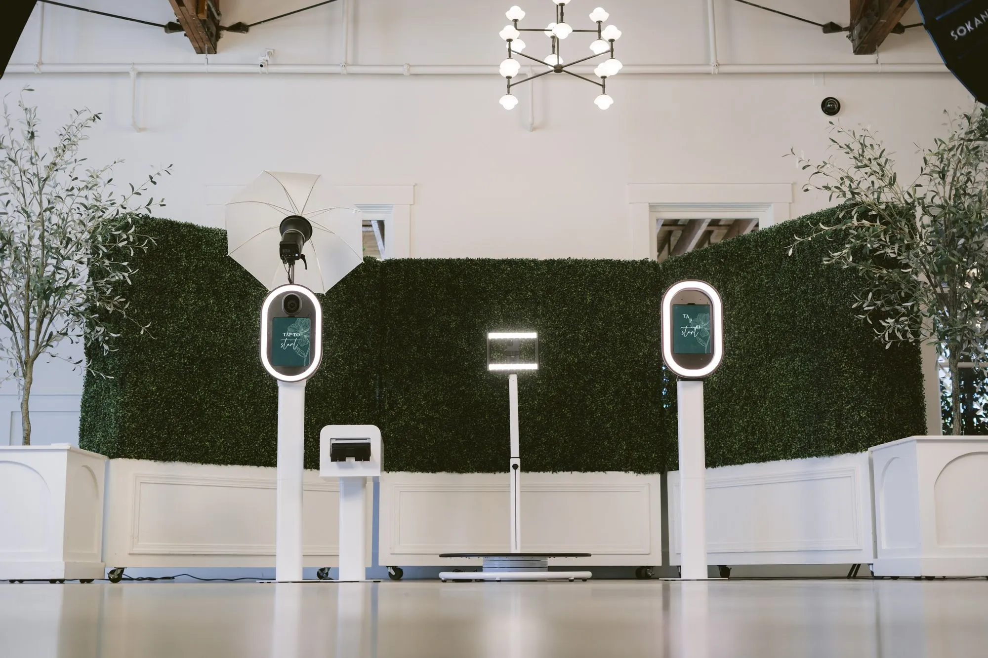 Photo booth hardware arranged in a bright event space with a hedge wall and chandelier