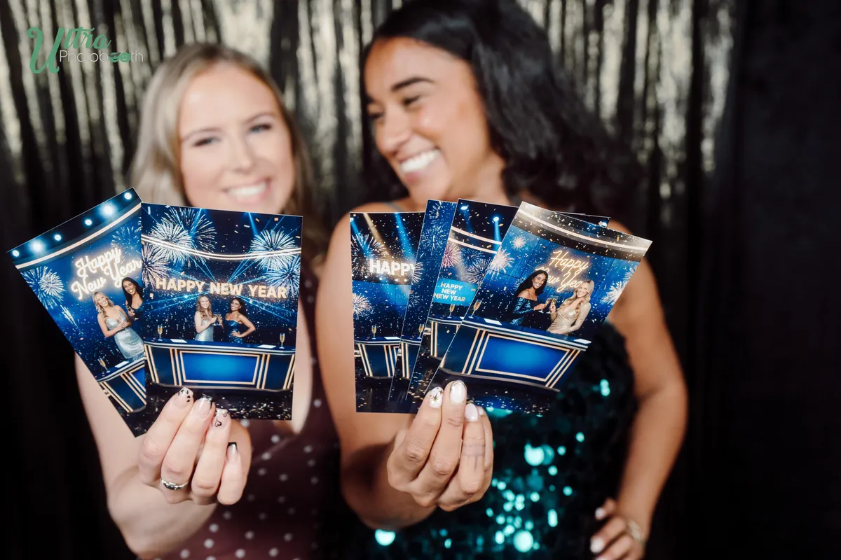 Two women holding blue Happy New Year photo prints in front of a dark metallic backdrop