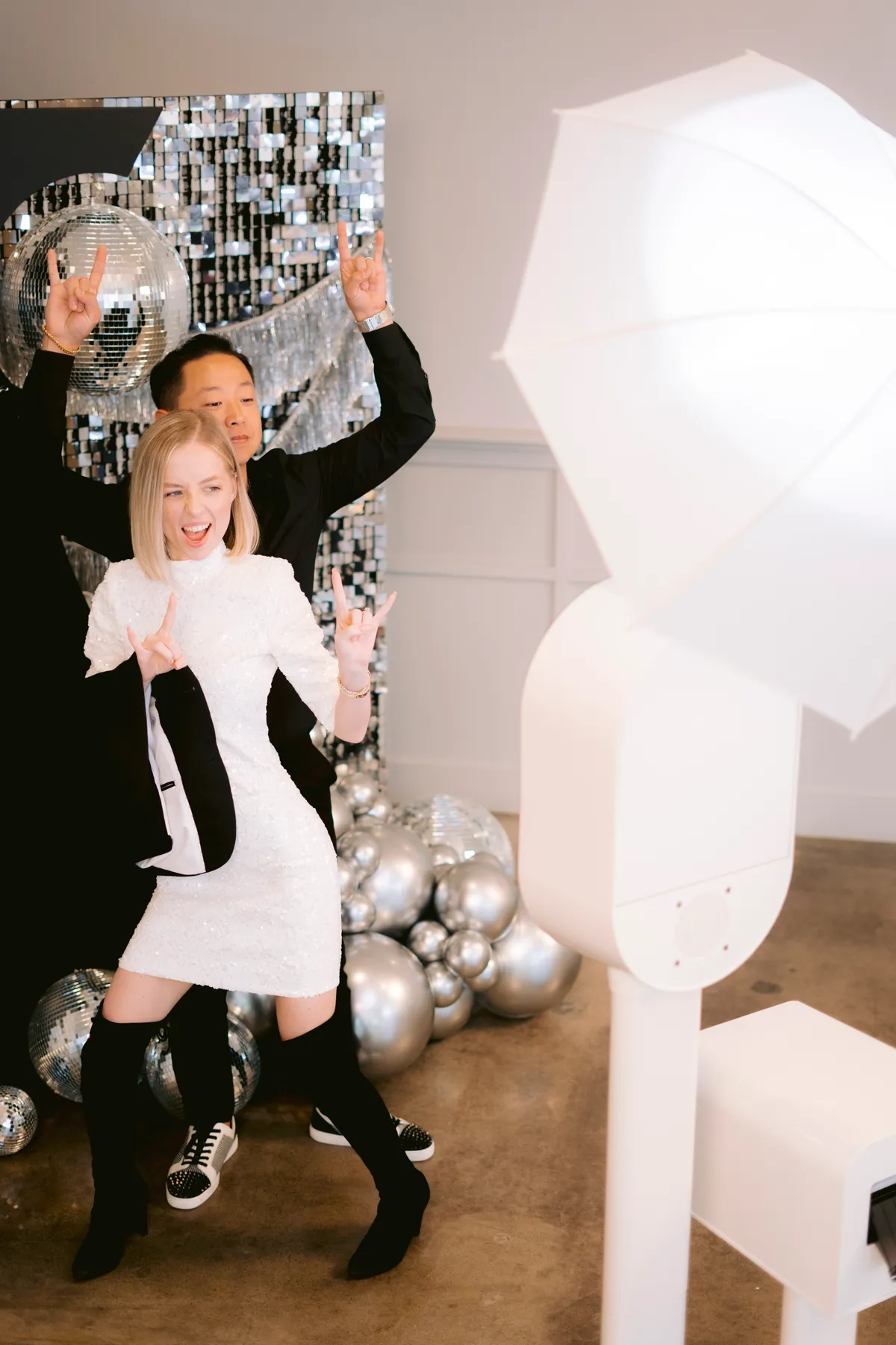 Two people posing beside a white photo booth, umbrella light, silver backdrop and disco balls
